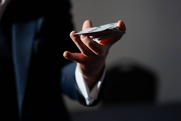 Stage illusion detail showing hand presenting stacked playing cards toward camera, selective focus on fingers and deck, minimal dark scene for magic theme