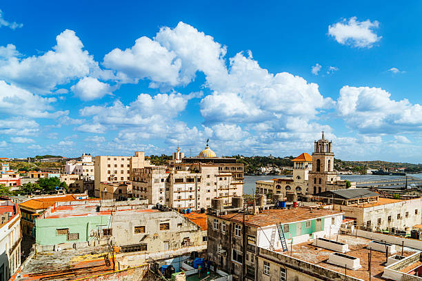 A vibrant Havana skyline reveals weathered buildings, colorful facades, terracotta rooftops, and a church tower along the waterfront. Bright blue sky and fluffy clouds convey warm, coastal colonial charm.