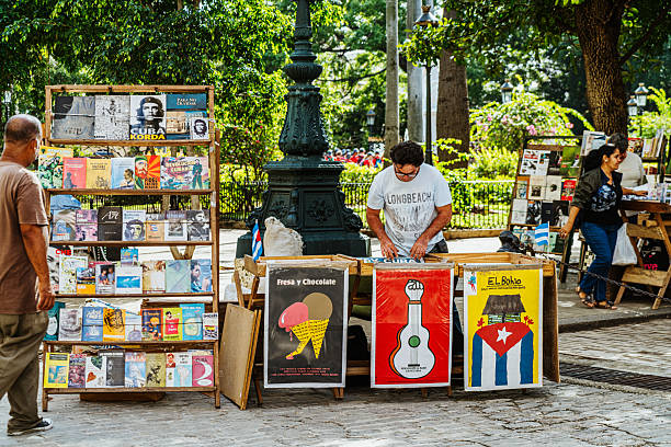 Havana, Cuba - February 23, 2017: Vibrant street market scene in Havana featuring display boards of posters, a man arranging prints, colorful artwork, and a Cubans flag-inspired poster; lively park setting with trees and pedestrians.
