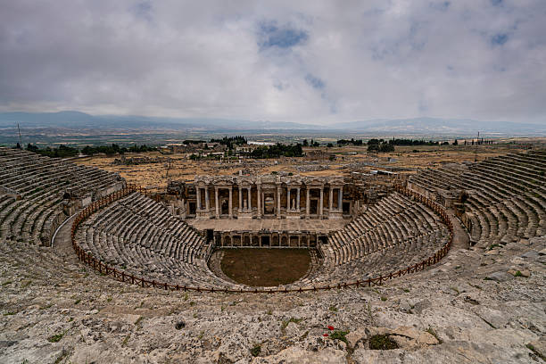 Dramatic sky frames panoramic view of ancient theater with complete semi-circular seating and stage building at Hierapolis, Turkey. The well-preserved structure shows the full extent of the Roman theater carved into the hillside with the ornate scaenae frons visible below. The monumental theater represents one of the best-preserved examples of Roman theatrical architecture in Turkey, demonstrating how Hierapolis combined therapeutic spa facilities with cultural amenities creating complete resort destination that attracted visitors from across the Roman world seeking both healing and entertainment.