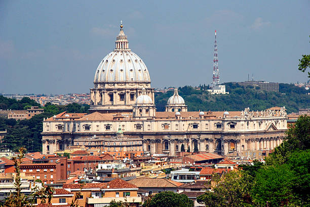 A high-angle, daytime view of the St. Peter’s Basilica facade and dome in Vatican City. The intricate Renaissance architecture is showcased against a backdrop of the Roman cityscape and a clear blue sky.