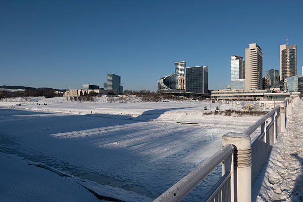 Modern skyline of Vilnius business district with frozen Neris River in winter. Snow-covered urban landscape featuring White Bridge and contemporary glass skyscrapers under clear blue sky in Lithuania.