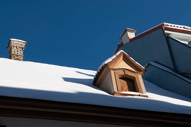Snow-covered historic rooftop with dormer window and chimney under a deep blue winter sky. Architectural detail of a traditional European building in cold weather, minimal composition with strong copy space. Ideal for winter, real estate, architecture, and seasonal design concepts.