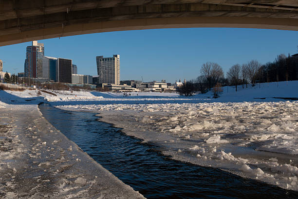 Winter cityscape of Vilnius, Lithuania, featuring the frozen Neris River and a modern urban skyline under clear blue sky. Snow-covered ice formations and contemporary architecture create a strong visual contrast, ideal for climate, winter weather, urban lifestyle, and European city concepts.