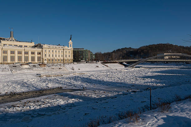 Winter cityscape of Vilnius, Lithuania, featuring the frozen Neris River and a modern urban skyline under clear blue sky. Snow-covered ice formations and contemporary architecture create a strong visual contrast, ideal for climate, winter weather, urban lifestyle, and European city concepts.