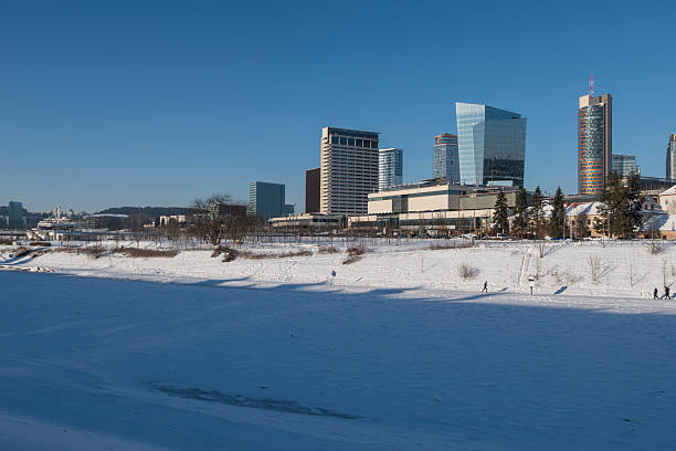 Winter cityscape of Vilnius, Lithuania, featuring the frozen Neris River and a modern urban skyline under clear blue sky. Snow-covered ice formations and contemporary architecture create a strong visual contrast, ideal for climate, winter weather, urban lifestyle, and European city concepts.