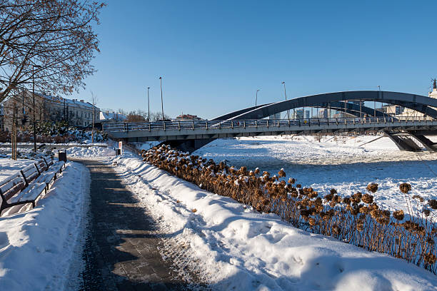 Winter cityscape of Vilnius, Lithuania, featuring the frozen Neris River and a modern urban skyline under clear blue sky. Snow-covered ice formations and contemporary architecture create a strong visual contrast, ideal for climate, winter weather, urban lifestyle, and European city concepts.