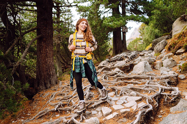 A young hiker walks along rocky hiking trail in a sparse forest. A smiling woman with colorful hiking backpack enjoys adventure and mountain scenery. Concept: hiking, recreation. Active lifestyle.