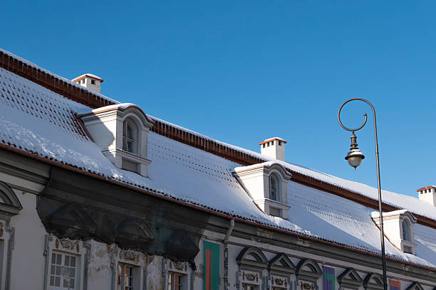 Winter scene of historic residential buildings in Vilnius Old Town, featuring snow-covered tiled rooftops, dormer windows, and a decorative street lamp against a clear blue sky. Classic European architecture with strong copy space, ideal for travel, seasonal, and urban concepts