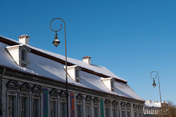 Winter scene of historic residential buildings in Vilnius Old Town, featuring snow-covered tiled rooftops, dormer windows, and a decorative street lamp against a clear blue sky. Classic European architecture with strong copy space, ideal for travel, seasonal, and urban concepts