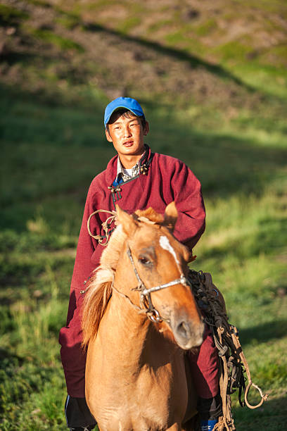 A young man wearing a maroon deel and blue cap sits astride a chestnut horse in a sunlit green valley. He holds the reins and looks toward the camera as rocky hills rise in the background, with warm late-afternoon light highlighting the landscape and the horse’s coat. The scene evokes pastoral, equestrian life and traditional rural clothing.