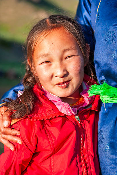 Close-up portrait of a young girl outdoors, smiling softly at the camera. She wears a bright red zip-up jacket over pink layers, her hair pulled into a braid. A hand rests gently on her shoulder and a blue embroidered garment with a bright green tassel is visible beside her. Warm golden light illuminates her face against a softly blurred natural background.
