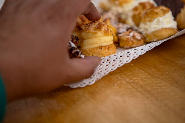 A hand with a delicate ring selects a cream puff from a generous assortment of golden pastries—including choux, shortbread, and chocolate-dusted treats—arranged on a lace paper tray.