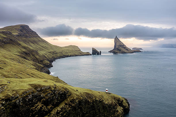 Dramatic coastal landscape featuring rugged green cliffs, the iconic Drangarnir sea stack and TindhÃ³lmur islet, and a small lighthouse