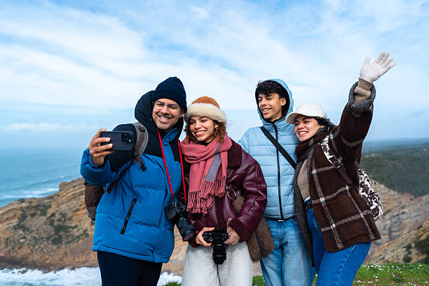 Group of diverse young adult friends smiling and posing for a camera while enjoying a sunset view over a city from a grassy hill, capturing joyful moments