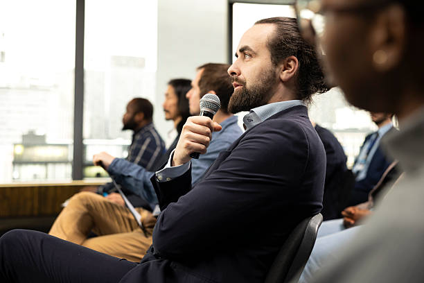 Seated male entrepreneur engaging in questions with microphone, interacting with speaker from the audience. International conference with audience engagement, business debate.