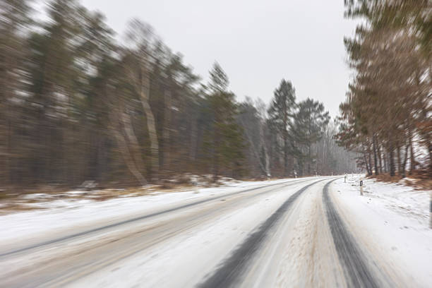 An empty snow covered road curves between trees on a cloudy day. Motion blur adds speed vibe and modern winter roadtrip mood.