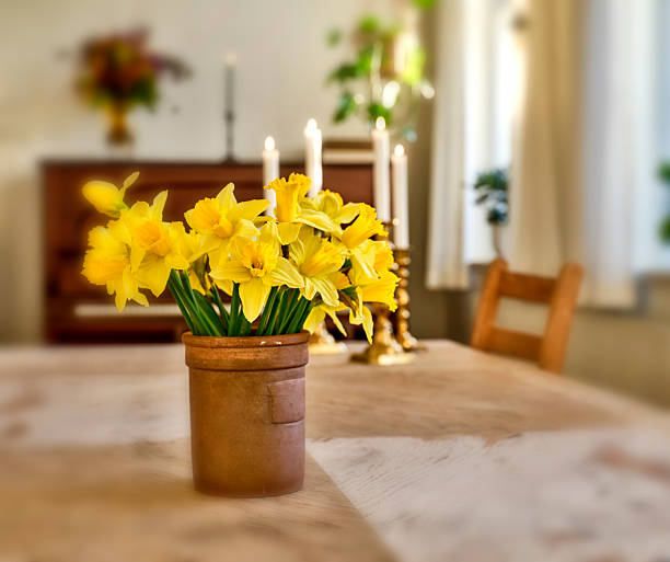 A cheerful bouquet of Narcissus daffodils in a rustic terracotta pot centers a wooden dining table. Soft candlelight and a blurred background hint at a calm, intimate home setting in Denmark, ideal for seasonal dÃ©cor, hospitality, and warm, welcoming interiors.