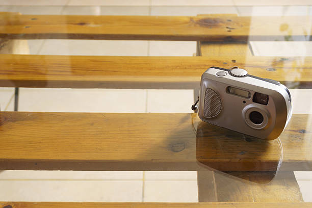 A silver compact 35mm film camera is positioned on a glass-topped table that reveals a light brown wooden slatted structure underneath, reflecting the bright ambient light of the room