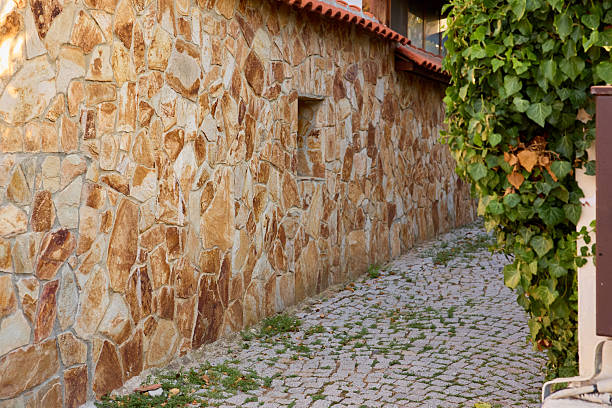 Rustic stone wall and cobblestone pathway beside green foliage and red-tiled roof.