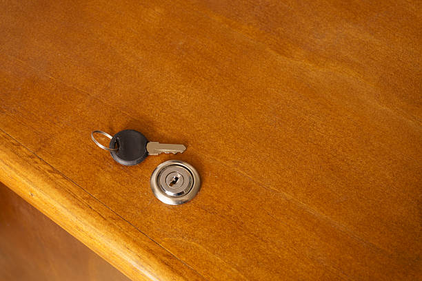 A close-up, top-down view of a single silver key with a black plastic head resting next to a circular metallic lock cylinder embedded in a smooth, light brown wooden cabinet or desk