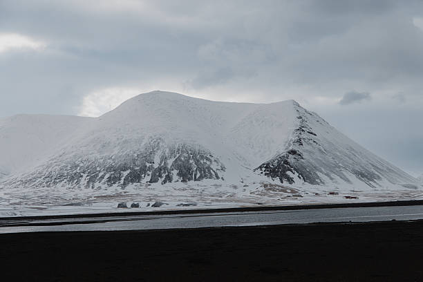 Snow-covered mountains rising above a dark, flat foreground and a body of water under a cloudy sky, showcasing the cold, rugged beauty of Arctic nature