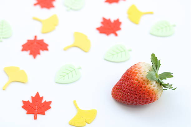 A ripe red strawberry surrounded by colorful autumn leaf decorations. This photo captures the beauty of seasonal fruit on a clean white background with a bright atmosphere.