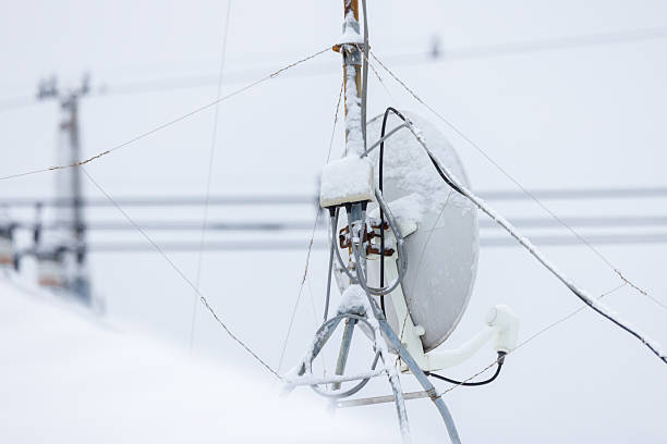 The back side of a satellite parabolic antenna (BS/CS dish) covered with snow due to snowfall. Frozen cables and metal fittings are visible. An image suggesting signal interference caused by severe winter weather conditions and maintenance of communication infrastructure in cold regions.
