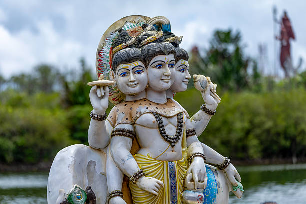 A vibrant, multi-faced statue of the Hindu Trimurti deity, adorned with intricate details and set in a serene natural landscape with water and trees