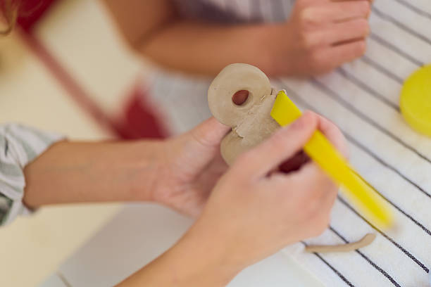 Two people gather around a striped tablecloth, shaping a clay figure with a yellow tool. This warm, collaborative moment captures creativity, learning, and shared project time in a home setting.