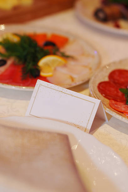 A table holds various dishes with vibrant food including fish, meats, and garnishes. A blank name card sits in front of an empty plate where food can be placed.