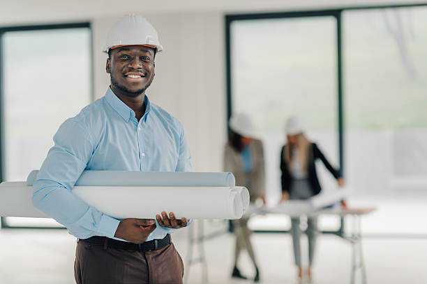 African american engineer proudly holding building plans, smiling at the camera, with colleagues working in the background