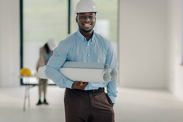 African american man, wearing a hard hat, standing in an empty room, holding rolled blueprints. Concept of architecture and construction