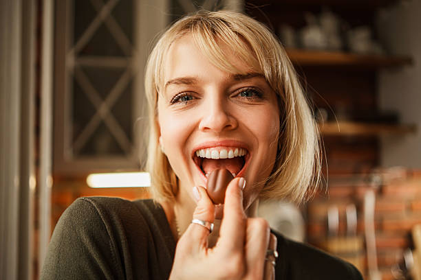 A cheerful blonde woman enjoys a chocolate praline while holding a plate of assorted chocolates. She is standing in a modern kitchen featuring white cabinetry and a rustic red brick wall.