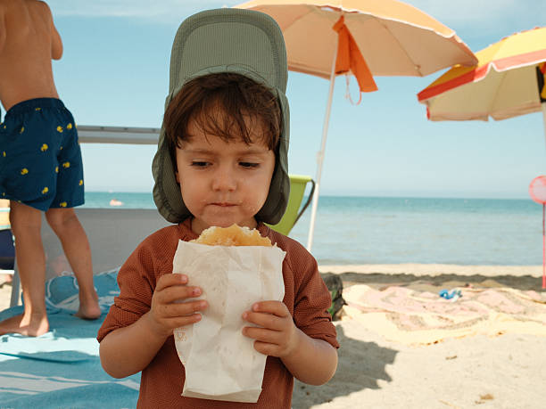 Young child enjoying a sweet treat on a sunny beach, wearing a hat for sun protection. Summer fun and childhood joy
