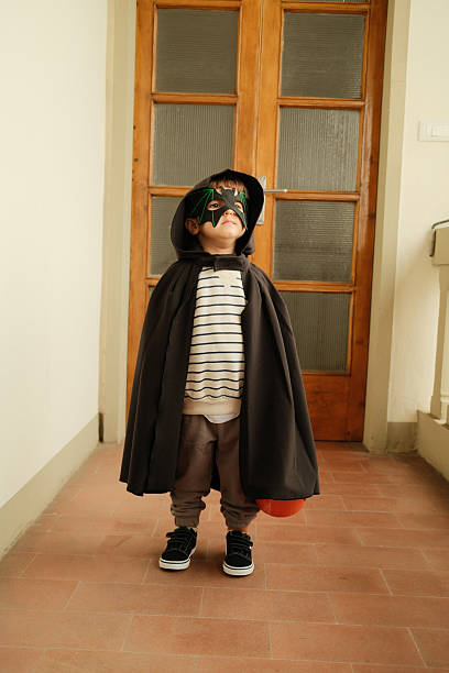 Little boy in bat costume with cape and mask, holding a candy bucket and looking up happily while trick-or-treating at home