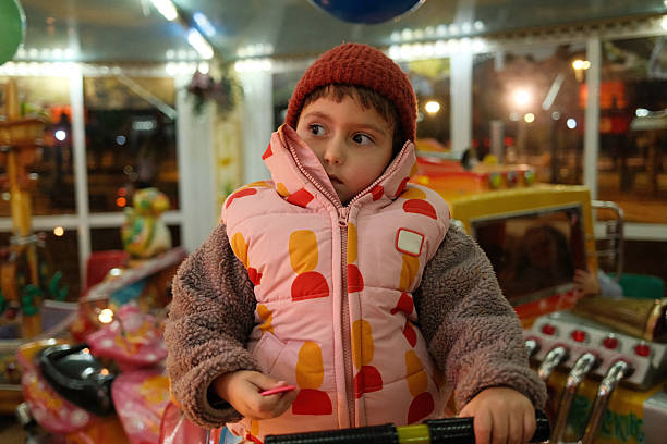 Young child wearing a hat and warm vest, sitting on a carousel ride at a festive amusement park at night