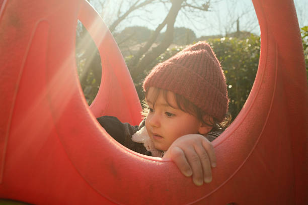 Young child wearing a red beanie, playfully peeking from a red slide on a sunny winter day. Childhood joy concept