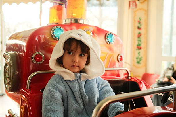 Young child wearing a hooded jumpsuit sitting in a red toy car at an amusement park enjoying childhood fun
