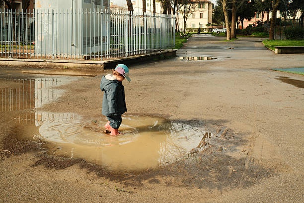 Young child wearing a hat and rubber boots stepping in a large muddy puddle in a park, creating splashes
