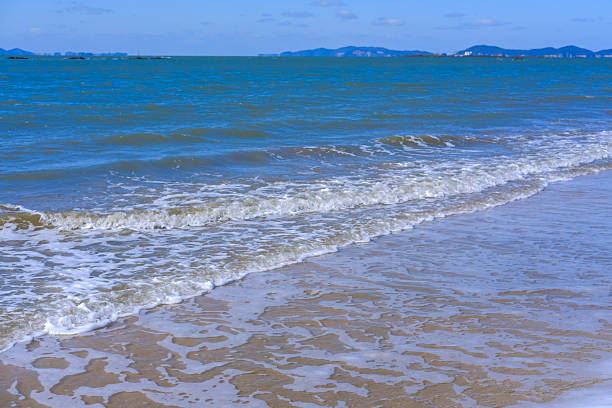 This image shows small waves rolling onto a sandy beach, with foamy water patterns in the foreground and calm blue sea stretching toward distant hills in South Korea.  The simple coastal scene is suitable for projects about relaxation, mindfulness, summer vacations, seaside travel, and peaceful ocean backgrounds in Asia.