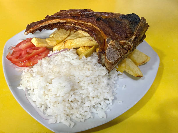 Close-up shot of a traditional Peruvian meal served on a white plate against a bright yellow background. A grilled beef chop rests over golden fries, paired with a generous portion of white rice and fresh sliced tomatoes with red onion. The simple presentation highlights everyday Peruvian comfort food and local dining culture.Concept:Peruvian cuisine, grilled beef chop, chuleta de res, arroz blanco, papas fritas, Latin American food, traditional meal, comfort food, South America cuisine, local restaurant dish, authentic flavors, simple plating, home-style cooking, chuleta de res a la plancha