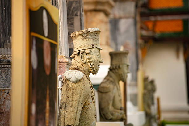 Profile view of an ancient stone guardian statue standing along the corridor of a historic Thai temple, captured with shallow depth of field to highlight intricate carving details and traditional architectural elements in soft natural light.
