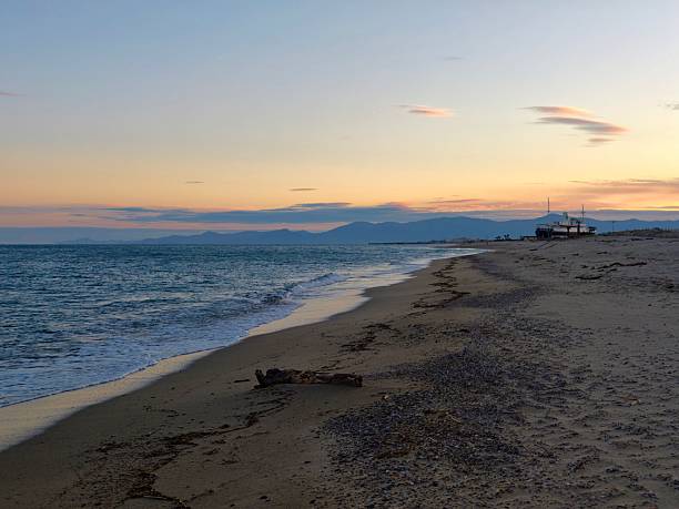 Sunset coastline with distant mountains and rolling surf, soft evening light across sand and water,