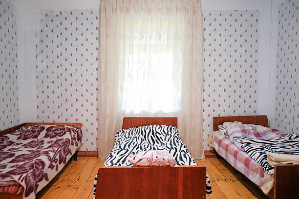 A simple and authentic bedroom interior featuring three wooden beds with colorful blankets. The central bed stands out with a bold black and white zebra pattern. Vintage floral wallpaper and natural wooden floors create a warm, nostalgic atmosphere for rustic accommodation themes.