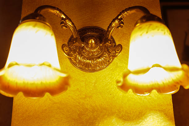 Warm vintage wall sconce casting glow. Ornate brass fixture with frosted glass shades mounted on textured plaster wall, bathing corridor in amber light closeup reveals filigree details, polished