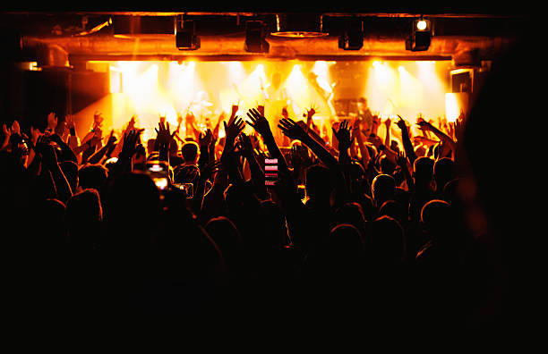Large audience with raised hands enjoys a live music concert under bright yellow stage lights in a club.