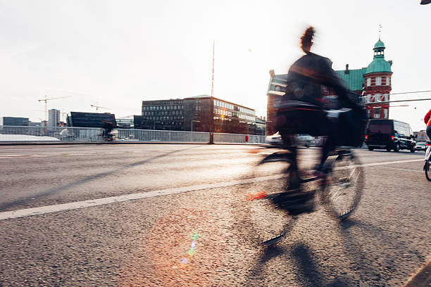Daily life unfolds in Copenhagen as people walk, cycle, and commute through the city. Motion blur emphasizes the rhythm of urban living, with pedestrians and cyclists moving past historic architecture, bridges, and streets. The scene reflects sustainable transport, active lifestyles, and the calm efficiency of a modern European capital where movement is constant but unhurried.