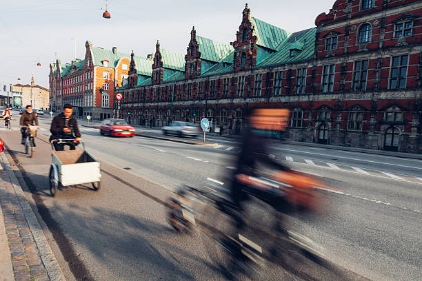 Daily life unfolds in Copenhagen as people walk, cycle, and commute through the city. Motion blur emphasizes the rhythm of urban living, with pedestrians and cyclists moving past historic architecture, bridges, and streets. The scene reflects sustainable transport, active lifestyles, and the calm efficiency of a modern European capital where movement is constant but unhurried.