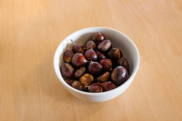 White ceramic bowl overflowing with glossy, brown chestnuts. The bowl is centered on a light, smooth wooden surface, creating a simple and clean aesthetic.